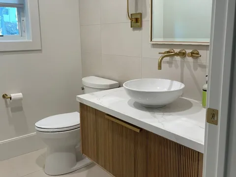 White-tiled bathroom with a fluted wood vanity, vessel sink, brass faucet, and toilet