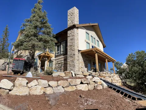Stone-clad home with tall chimney, evergreen tree, and landscaped rock wall